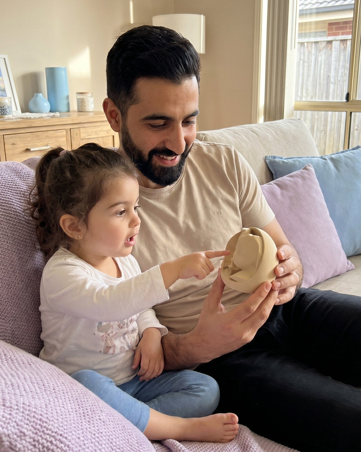 A father and daughter sitting on a couch in a living room, looking at and holding a 3D print keepsake, which represents her size and development when she was in her mother's womb.
