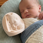 A sleeping newborn baby is swaddled in a cream and blue blanket, with a beige 3D print keepsake resting beside the baby, which is a replica of the baby when they were in the womb.